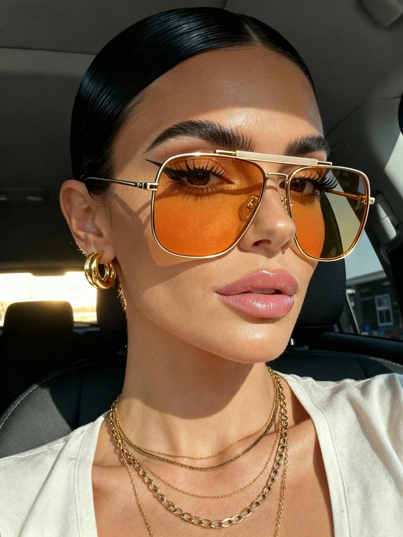 Woman wearing gold sunglasses and layered necklaces inside a car.