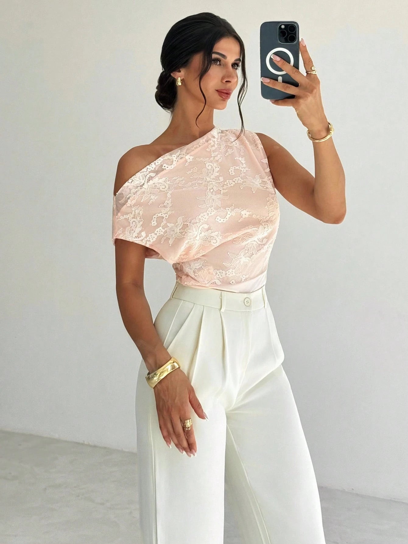 Woman taking a mirror selfie wearing a pink lace top and white pants.