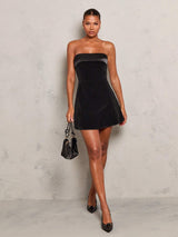 Woman wearing a black strapless dress holding a matching handbag against a gray background