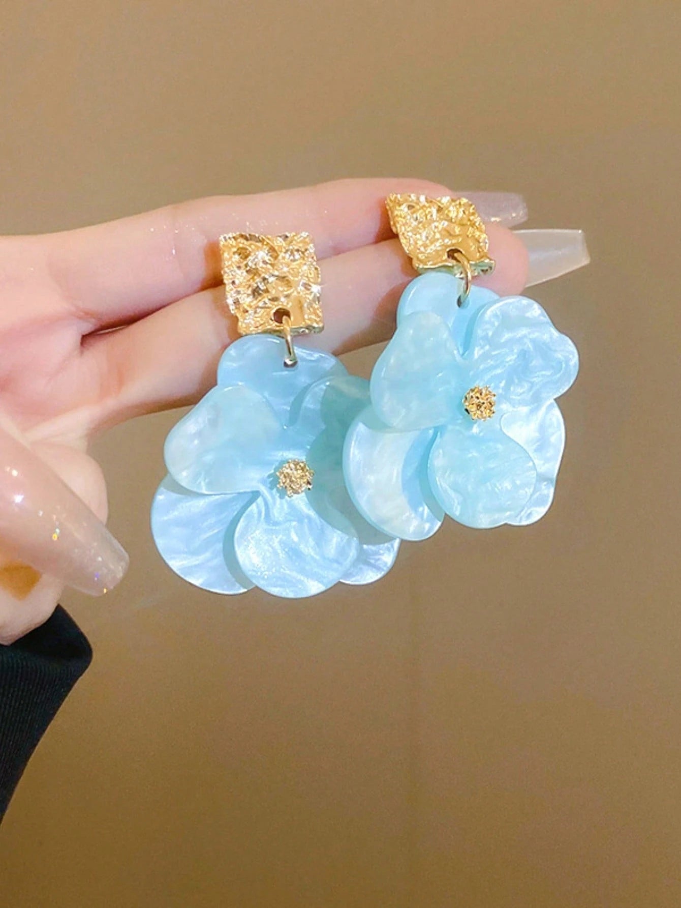 Light blue floral earrings with gold accents held by a hand against a beige background