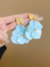 Light blue floral earrings with gold accents held by a hand against a beige background