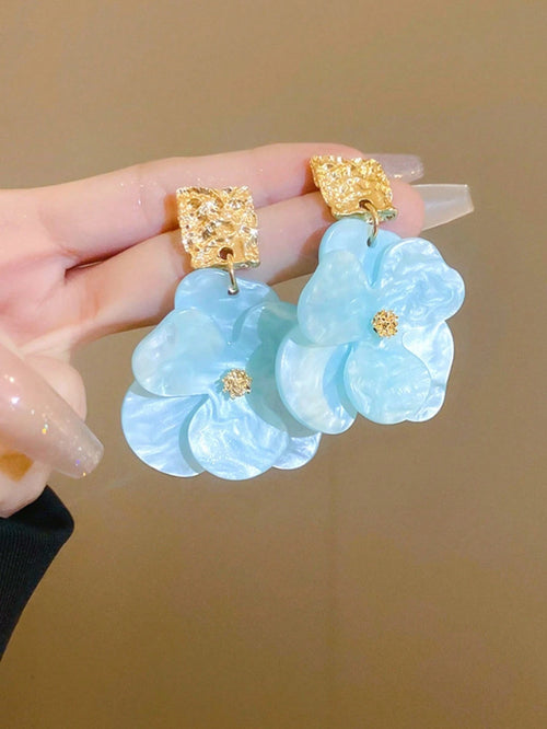 Light blue floral earrings with gold accents held by a hand against a beige background