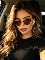 A woman with long wavy blonde hair wearing cat-eye sunglasses with gold frames and brown lenses, sitting in a car. She is wearing a black t-shirt and white wireless earbuds.