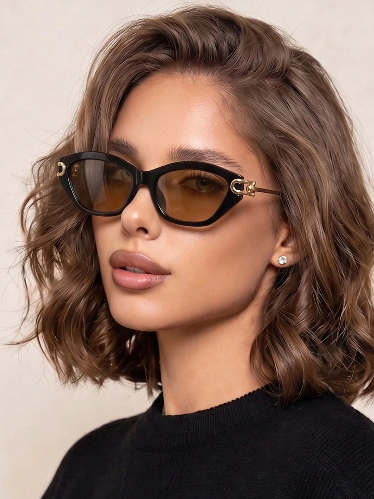 A close-up of a woman with wavy brown hair wearing black cat-eye sunglasses with gold-tone metal accents and brown-tinted lenses, paired with a black top against a light beige background.