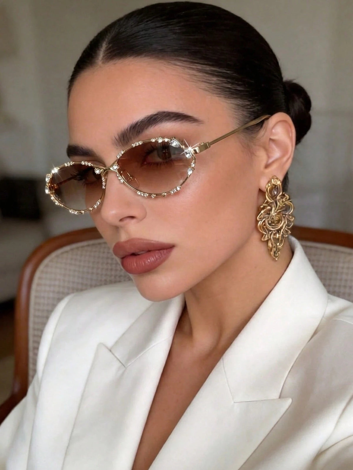 A close-up of a woman wearing oval sunglasses with rhinestone-trimmed gold frames and brown lenses, paired with a white blazer and ornate gold earrings.