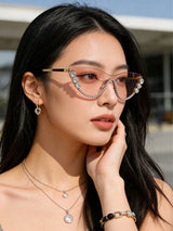 A woman wearing stylish cat-eye sunglasses with rhinestone embellishments on the rims, posed outdoors with her hand near her face.