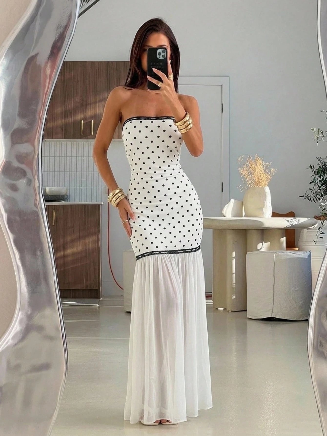 A woman takes a mirror selfie wearing a strapless white maxi dress with black polka dots on the bodice and a flowing white skirt. The setting is a modern, minimalist interior with neutral tones.