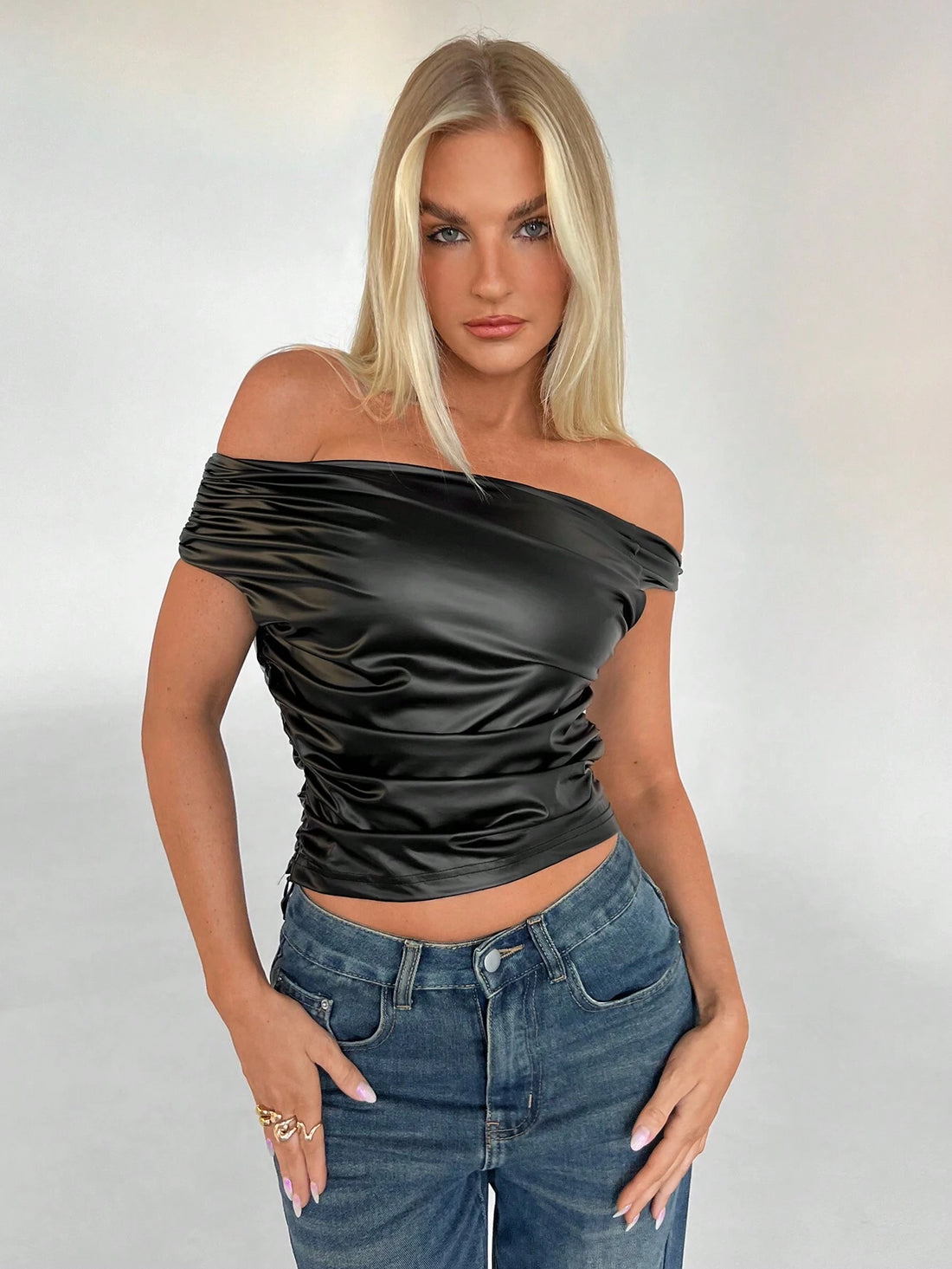 A blonde model wearing a black off-the-shoulder ruched satin crop top and blue jeans, standing against a light gray studio background.