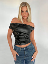 A blonde model wearing a black off-the-shoulder ruched satin crop top and blue jeans, standing against a light gray studio background.