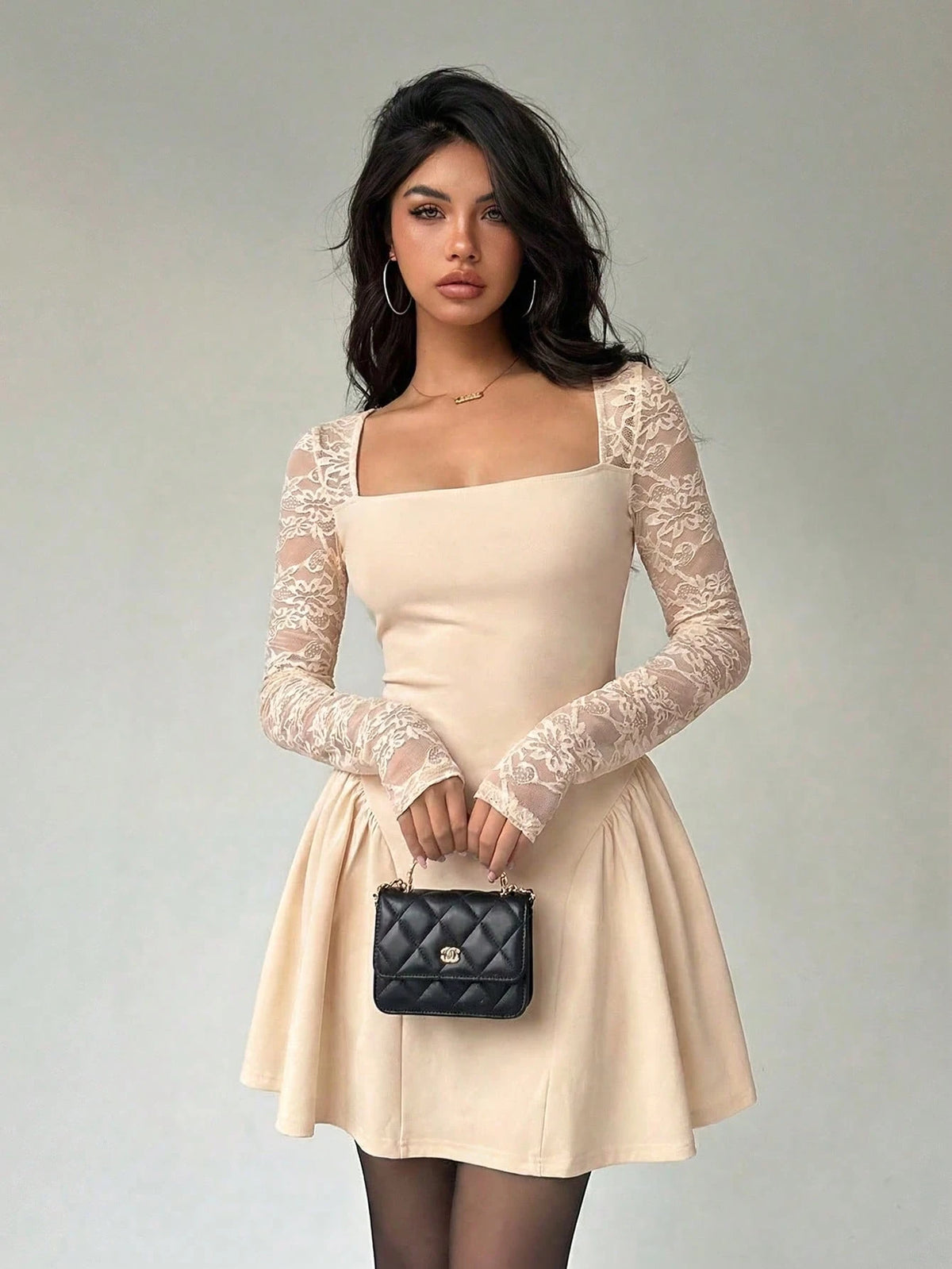 A woman wearing a beige mini dress with long sheer lace sleeves, a square neckline, and a flared skirt, shown against a plain light gray studio background.