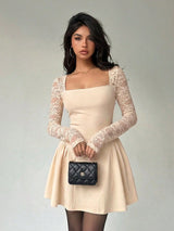 A woman wearing a beige mini dress with long sheer lace sleeves, a square neckline, and a flared skirt, shown against a plain light gray studio background.