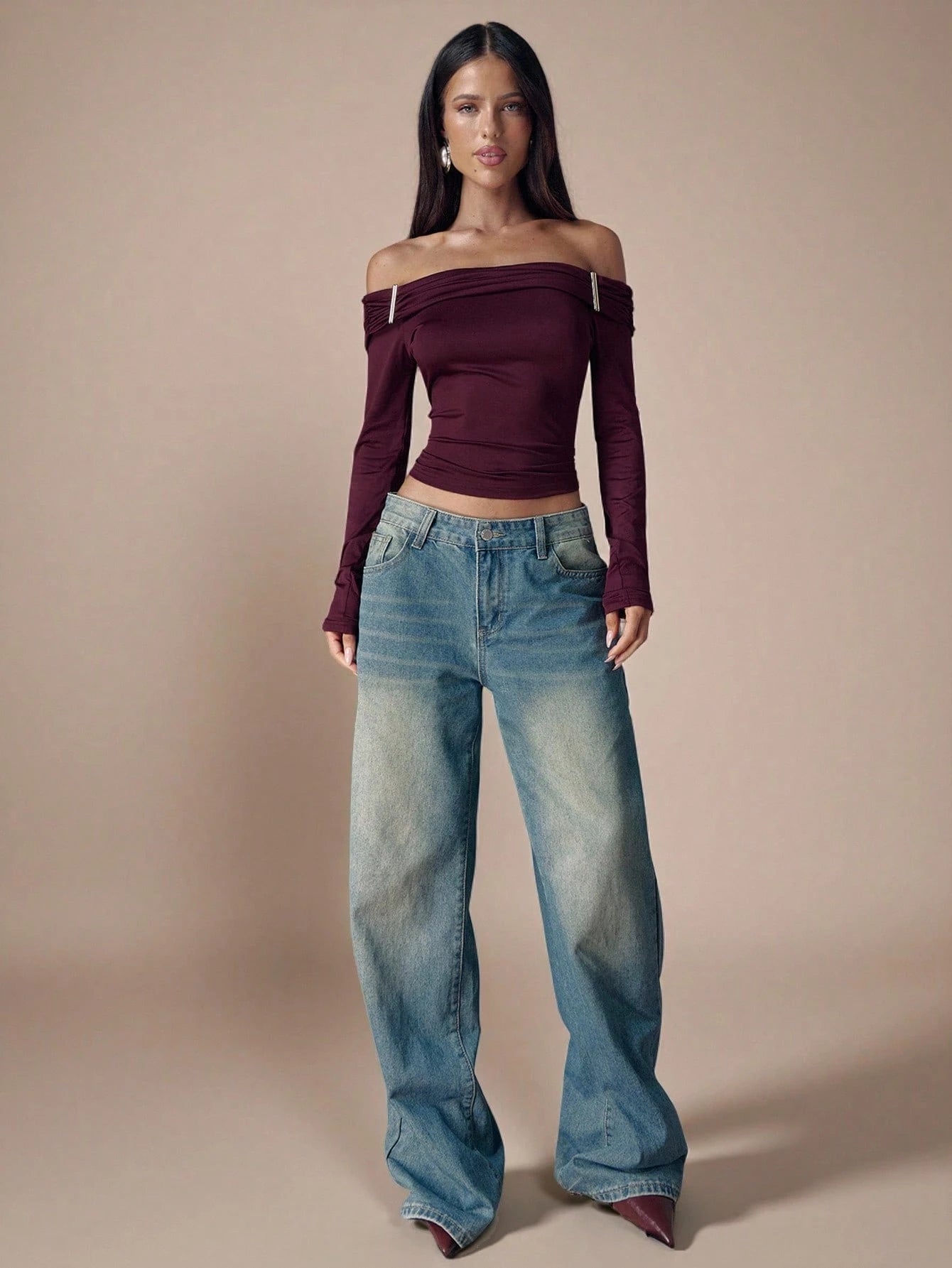Woman wearing a burgundy off-shoulder top and blue jeans against a beige background