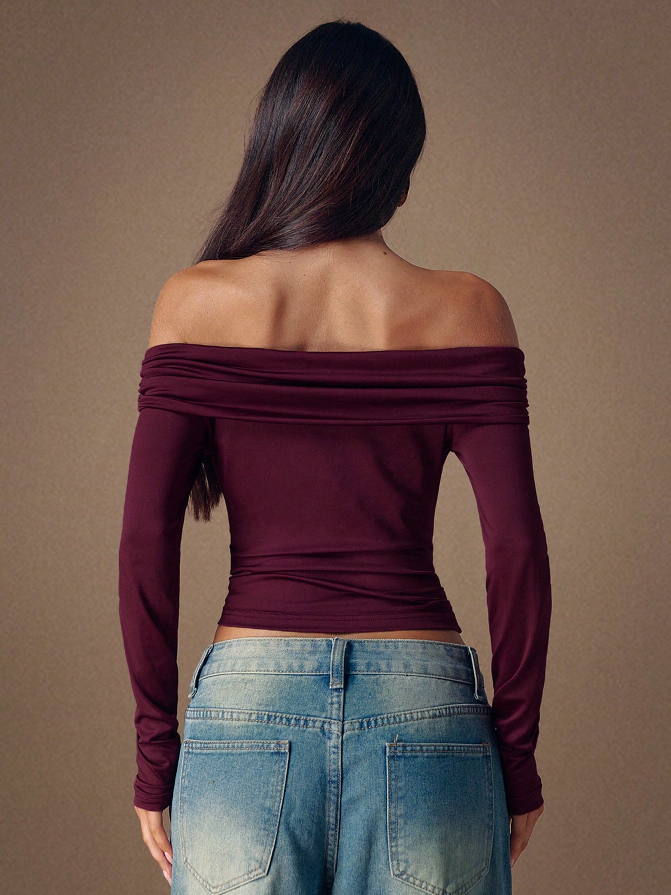 Person wearing a burgundy off-shoulder top and blue jeans against a brown background