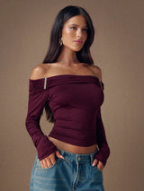 Woman wearing a burgundy off-shoulder top and blue jeans against a brown background