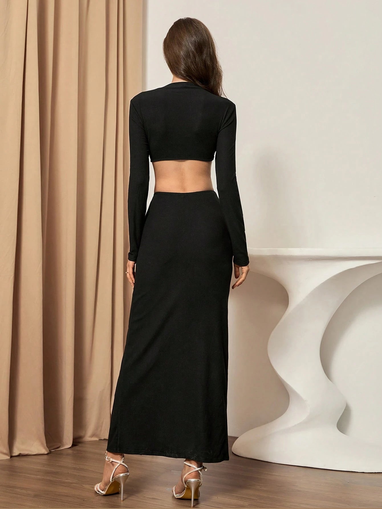 Woman wearing a black long-sleeve top and black skirt standing in front of a beige curtain.