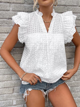 A woman wearing a white textured short-sleeve blouse with ruffled shoulders and a split neckline, paired with denim shorts, standing against a gray brick wall.