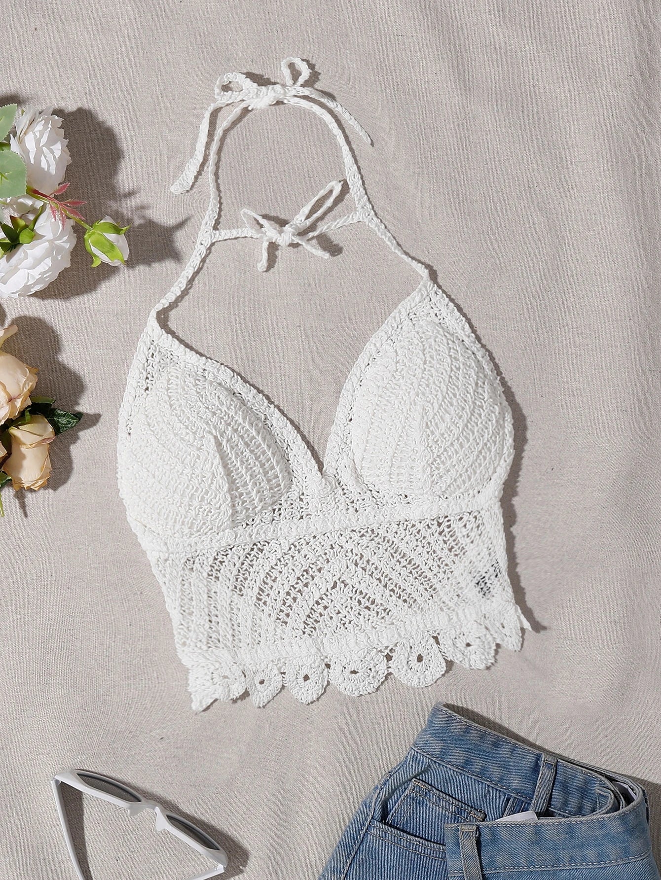 White crochet halter bikini top laid flat on a beige fabric background, styled with sunglasses, flowers, and folded blue jeans.