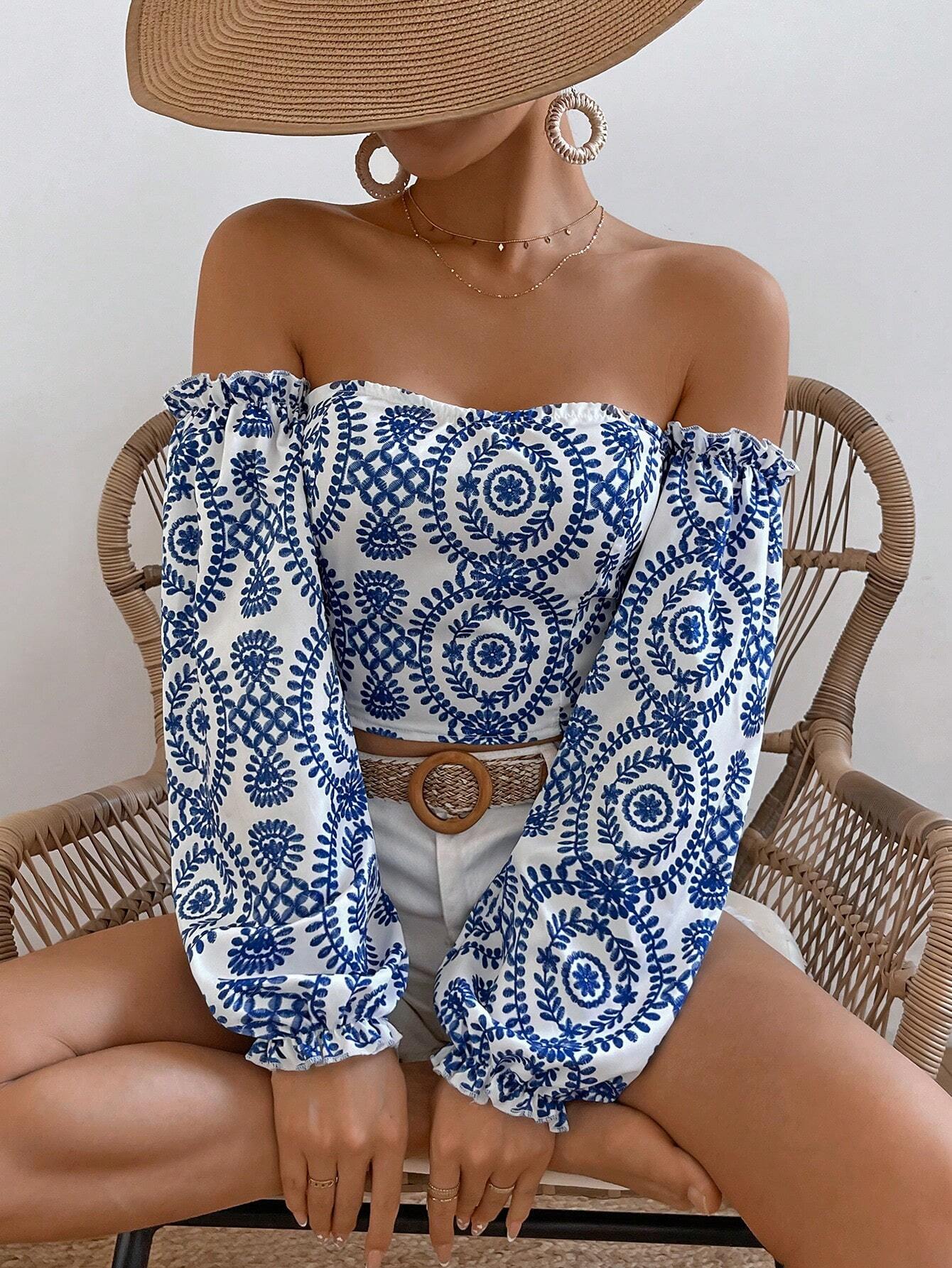 A woman seated in a wicker chair wearing an off-the-shoulder long-sleeve top with a blue and white circular floral pattern, paired with white shorts and a brown belt.