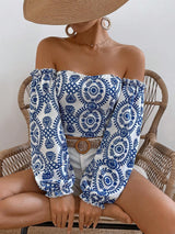 A woman seated in a wicker chair wearing an off-the-shoulder long-sleeve top with a blue and white circular floral pattern, paired with white shorts and a brown belt.