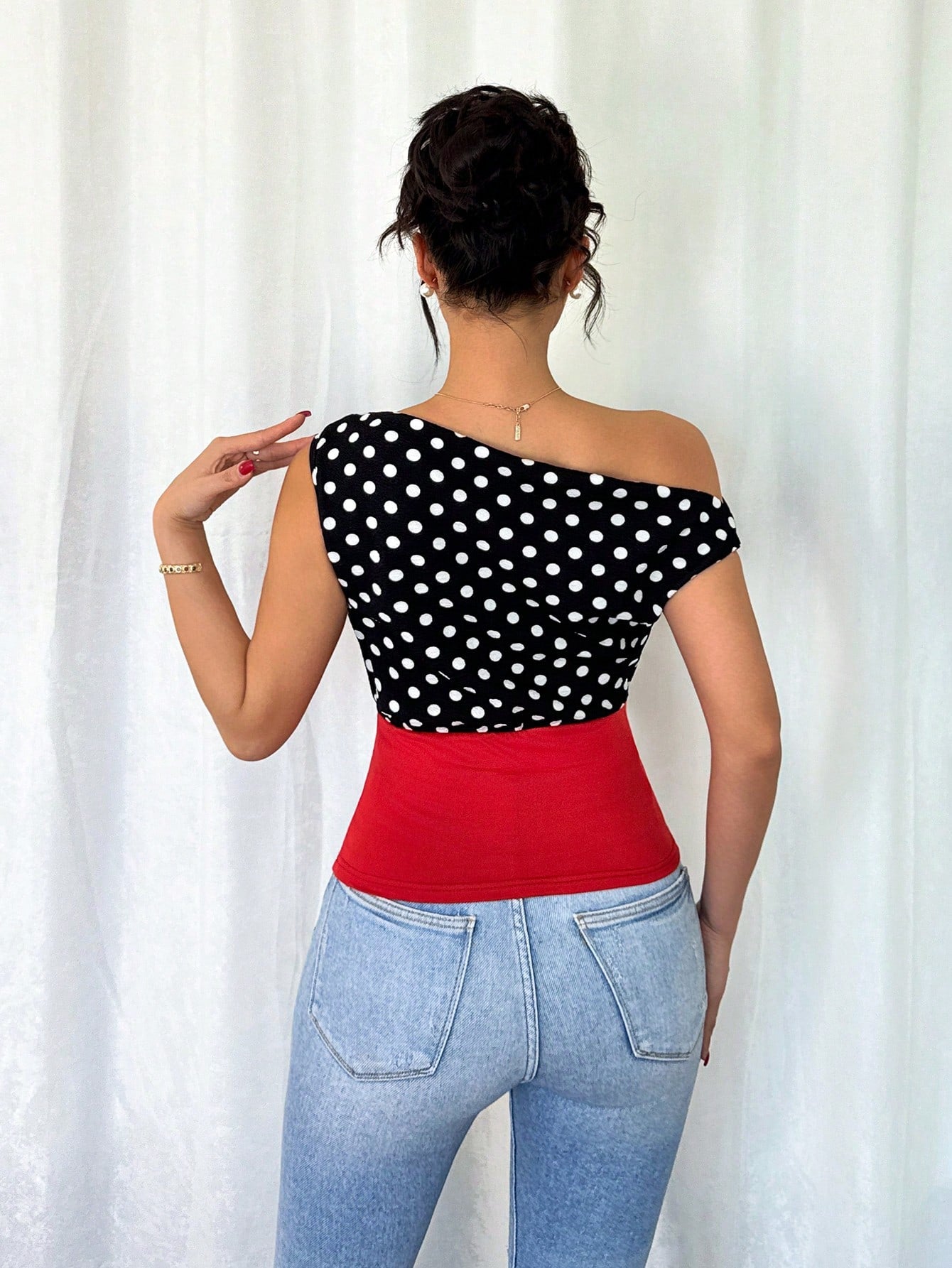 Woman wearing a black polka dot top and red waistband with blue jeans against a white curtain background