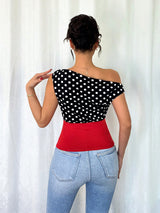 Woman wearing a black polka dot top and red waistband with blue jeans against a white curtain background