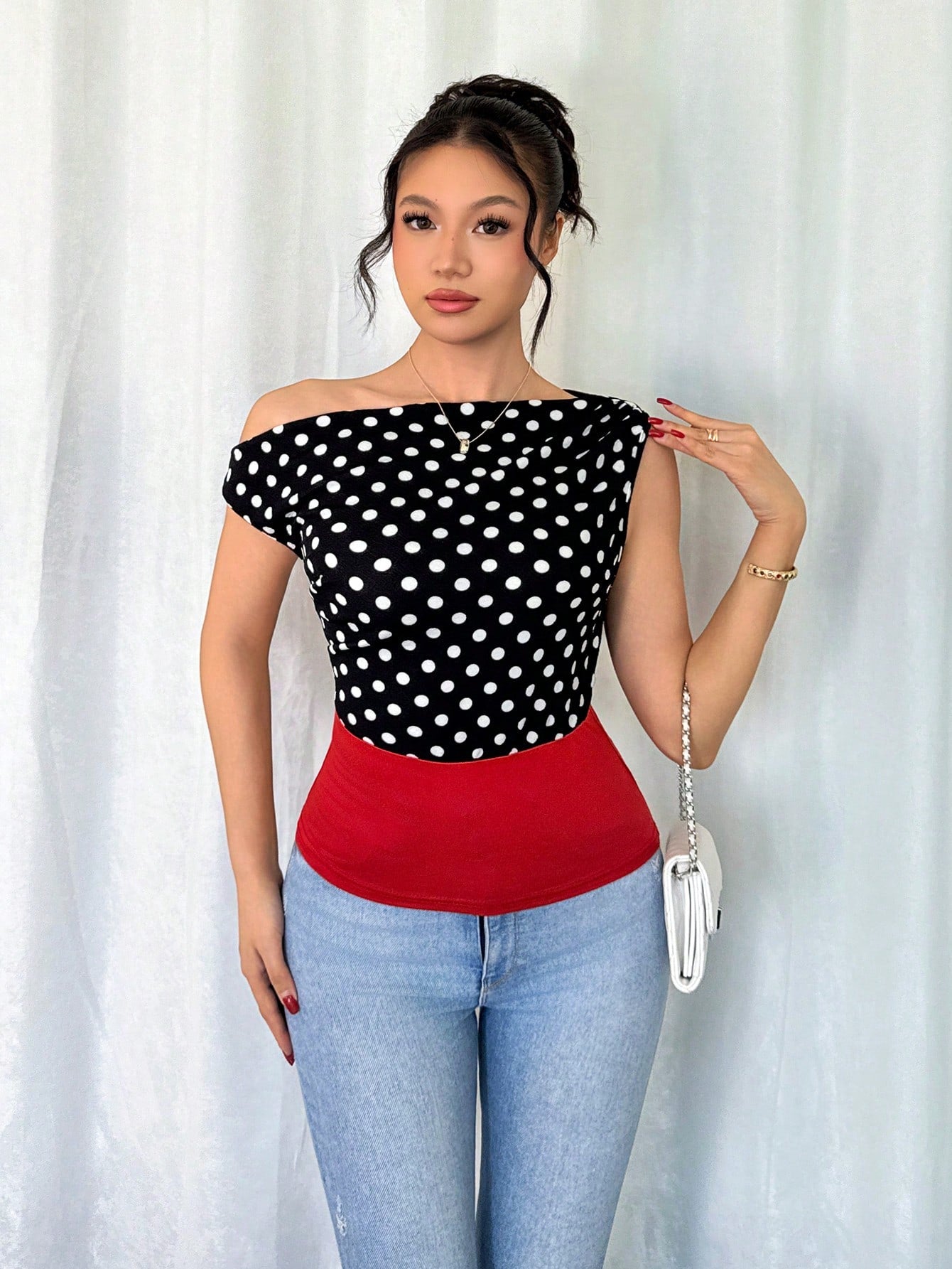 Woman wearing a black and white polka dot top with a red bottom and blue jeans, standing against a white curtain.