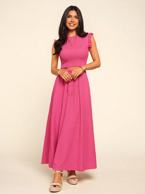 A woman modeling a bright pink two-piece set consisting of a sleeveless ruffle-shoulder crop top and a matching long skirt, paired with neutral platform sandals against a solid cream background.