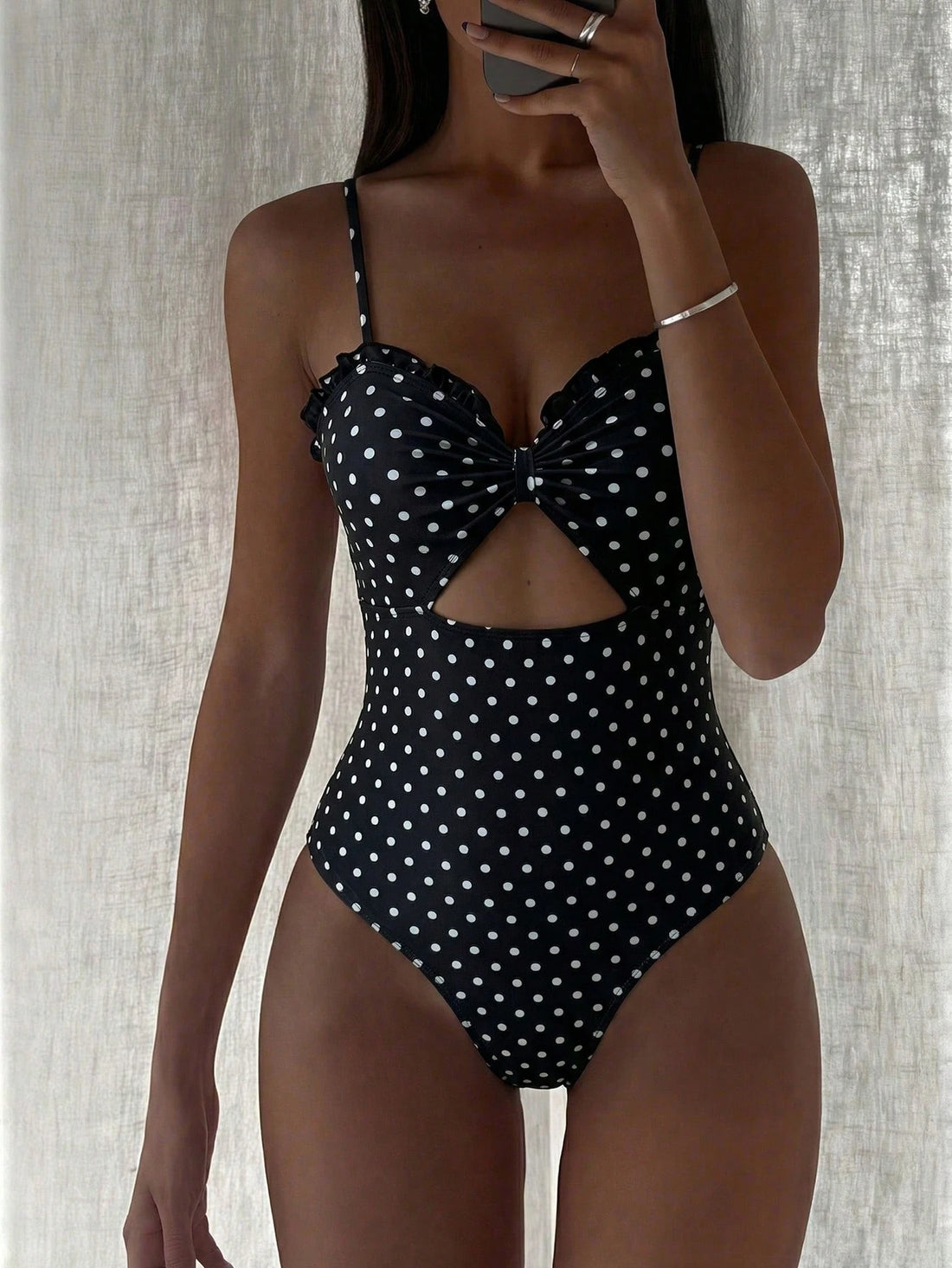 A woman modeling a one-piece swimsuit with a black and white polka dot pattern, featuring a sweetheart neckline, thin shoulder straps, a central cutout, and a large bow detail at the bust. She is holding a phone in front of her face against a textured light gray curtain background.