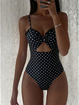 A woman modeling a one-piece swimsuit with a black and white polka dot pattern, featuring a sweetheart neckline, thin shoulder straps, a central cutout, and a large bow detail at the bust. She is holding a phone in front of her face against a textured light gray curtain background.
