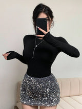 A woman takes a mirror selfie wearing a fitted black long-sleeve top and a silver sequined mini skirt, with a black phone covering part of her face. A beige sofa is visible in the background.