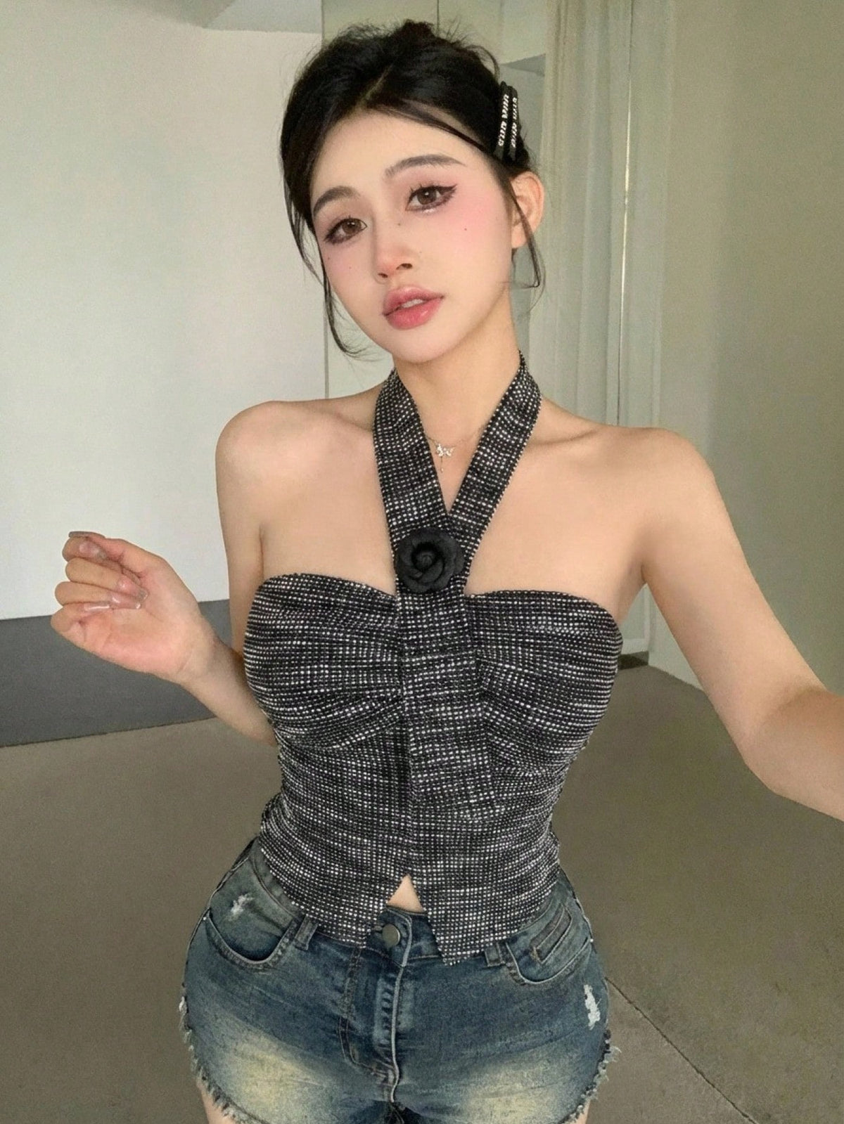 A woman modeling a black and gray textured halter crop top with a large black flower detail at the center of the neckline, paired with distressed denim shorts.