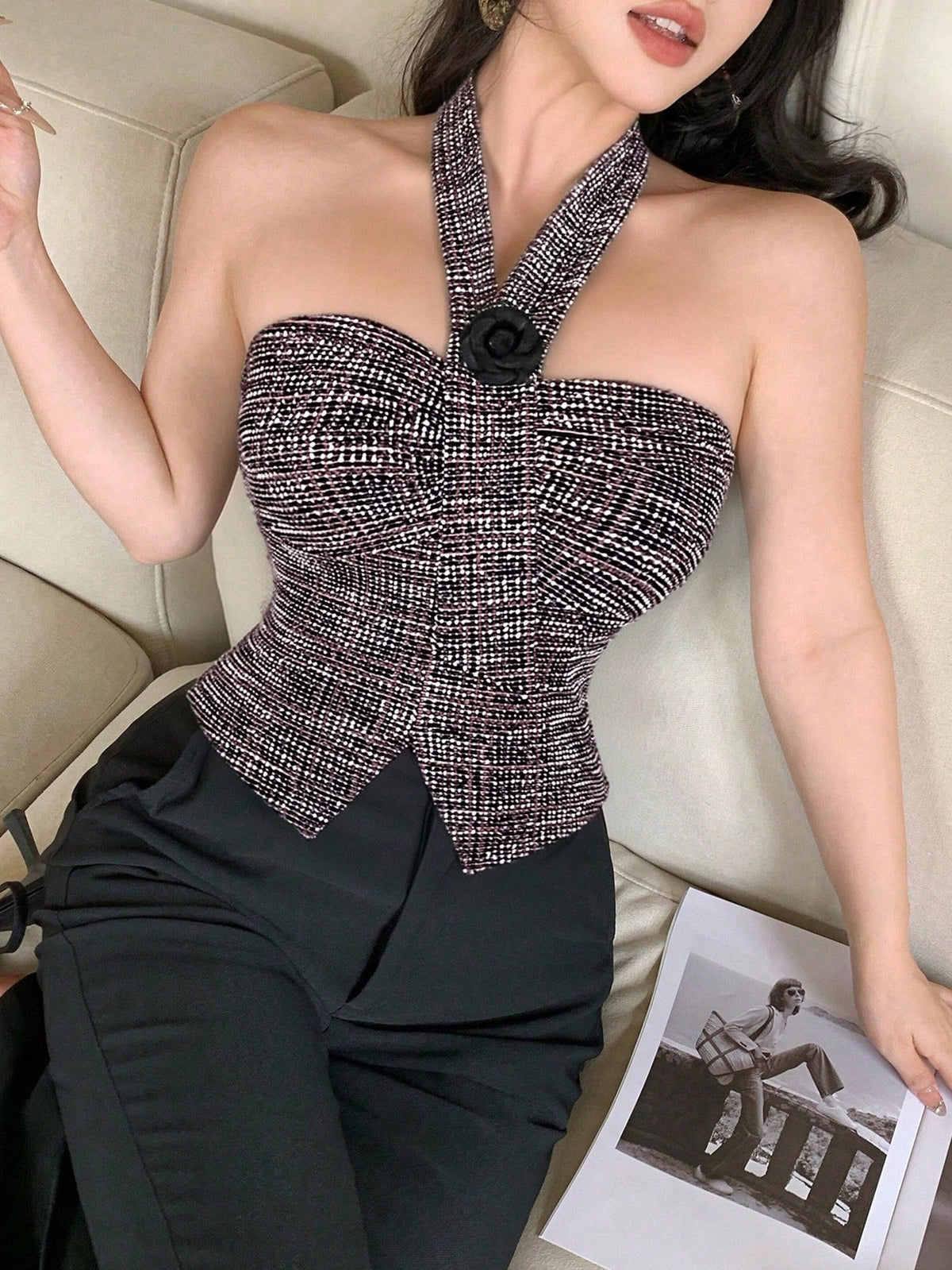 A woman wearing a strapless halter-style top with a textured black and beige tweed pattern, a black rose detail at the neckline, and black pants, posed on a beige sofa with a magazine nearby.