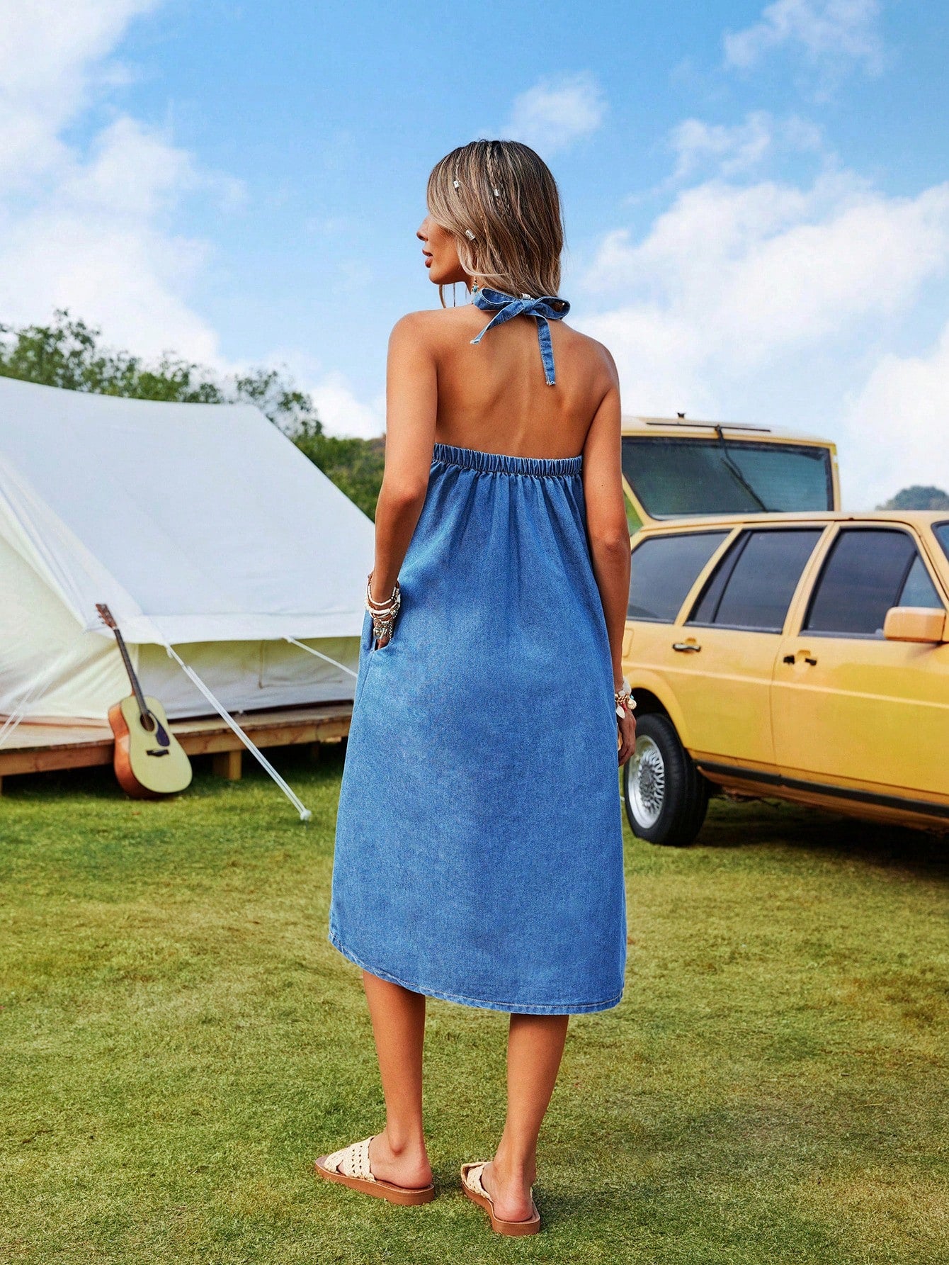 Woman in a blue dress standing in a grassy area with a tent and yellow car in the background.