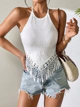A woman wearing a white sleeveless halter top with a textured sunburst pattern and a beaded fringe hem, paired with light-wash denim shorts and carrying a woven straw shoulder bag.