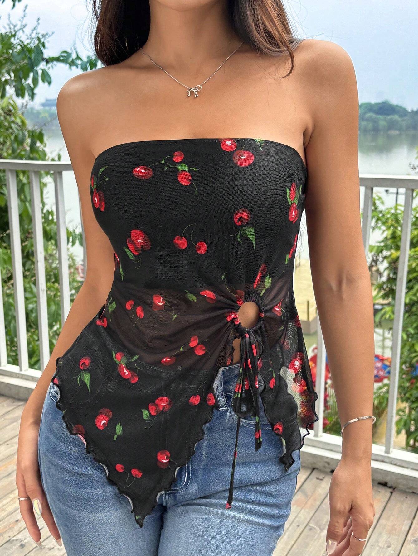 A woman wearing a black strapless top with a red cherry print and a side tie detail, paired with blue jeans, standing on a wooden deck with a railing and water in the background.