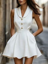 A woman wearing a sleeveless white mini dress with a structured collar, three gold-tone decorative buttons down the front, a matching waistband, and a flared skirt, photographed outdoors on a blurred street background.