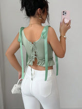 Woman wearing a green top with ribbon straps and white pants, taking a mirror selfie.
