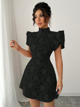 A woman modeling a black short-sleeve mini dress with a high mock neck and textured floral jacquard pattern, standing in front of a white curtain and wall.