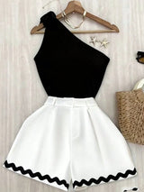 A women's two-piece outfit consisting of a black one-shoulder top and white high-waisted shorts with a black wavy trim, displayed on a wooden hanger against a light wood background.