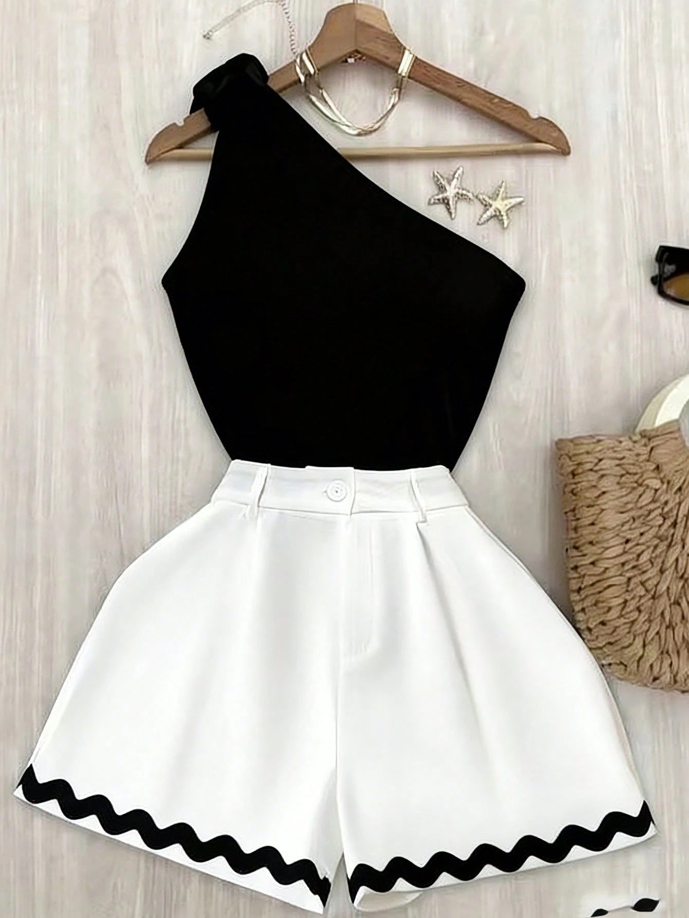 A women's two-piece outfit consisting of a black one-shoulder top and white high-waisted shorts with a black wavy trim, displayed on a wooden hanger against a light wood background.