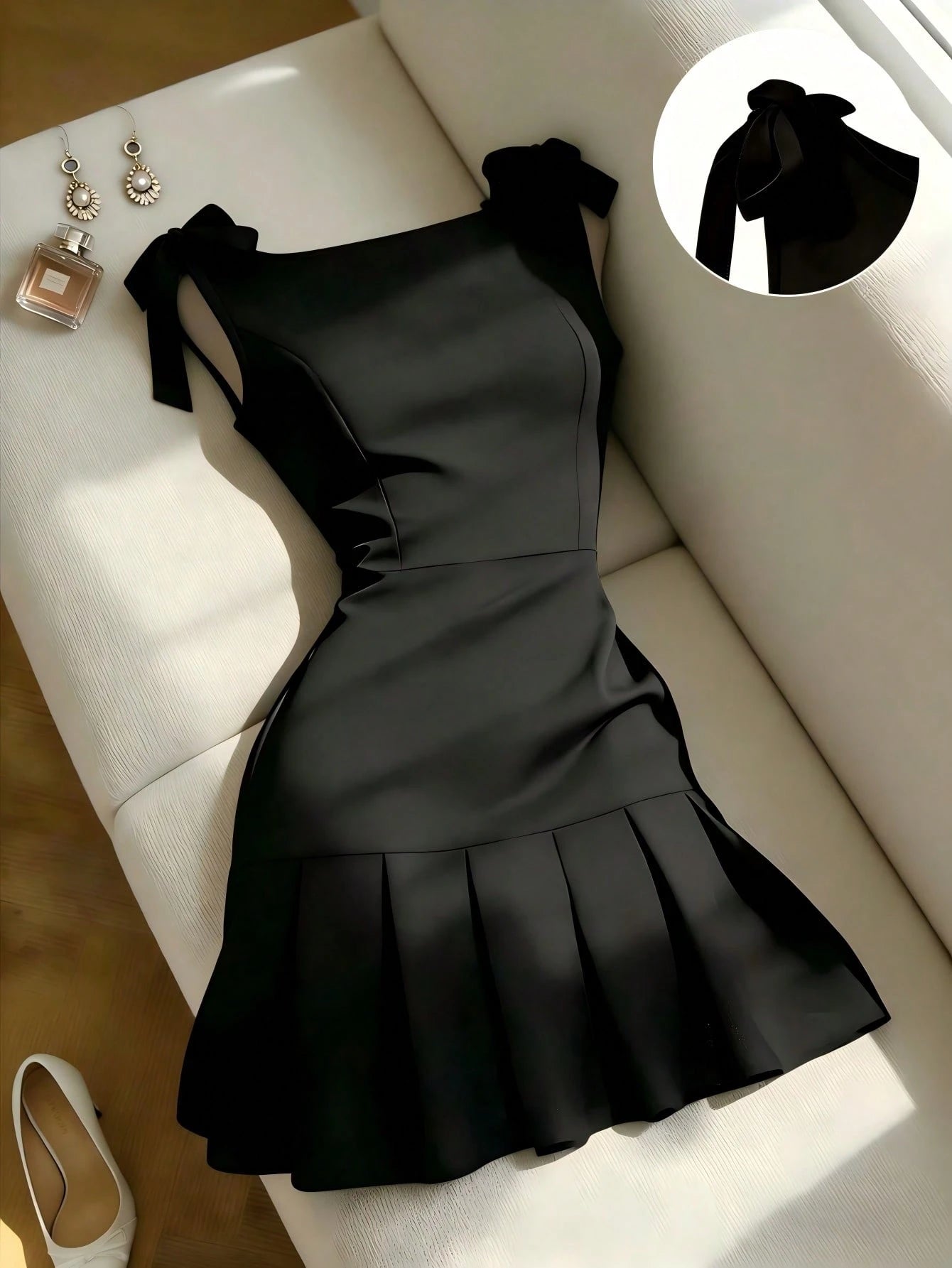 Black dress on a white surface with a close-up of a bow detail, earrings, and a perfume bottle.