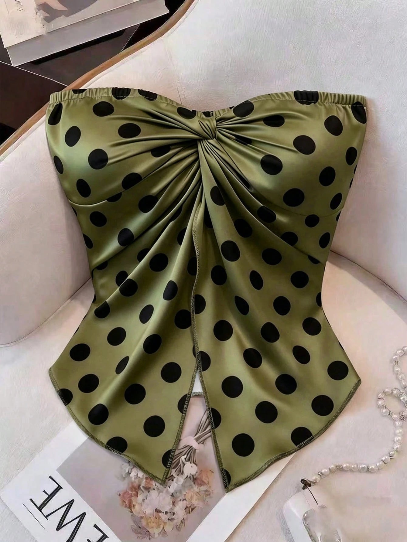 An olive green satin bandeau top with black polka dots and a large front bow, styled on a cream-colored armchair with a pearl necklace and magazine nearby.