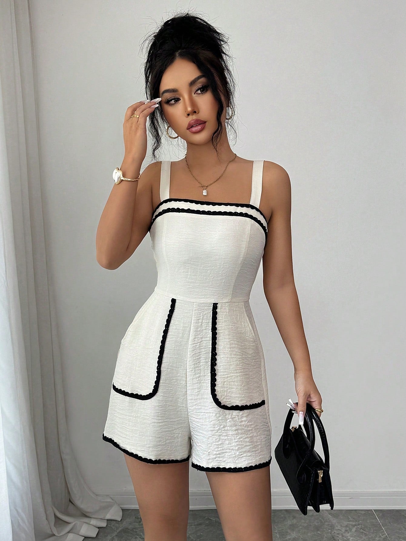 A woman modeling a white textured romper with black trim details, holding a small black handbag against a neutral indoor background.