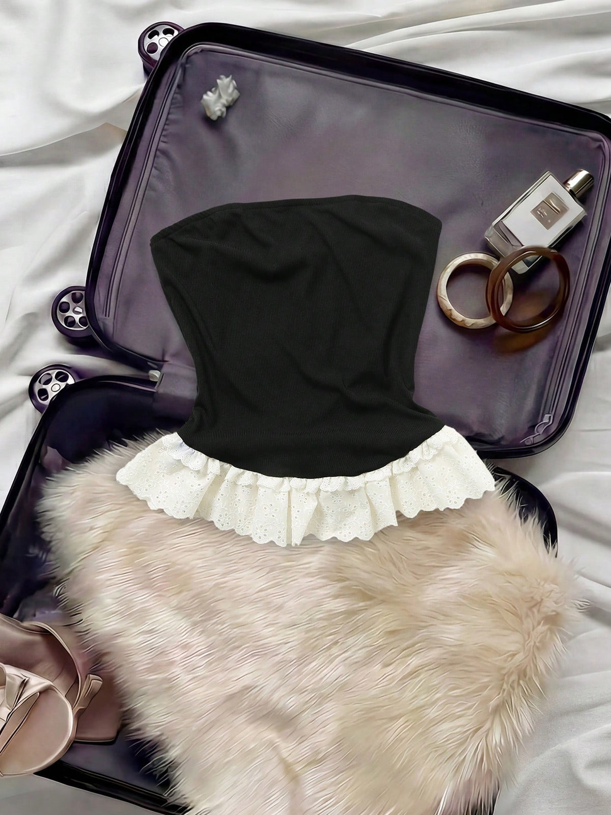 A black strapless tube top with a white ruffled lace hem, styled inside an open black suitcase with a beige faux fur accessory, rings, and a perfume bottle.