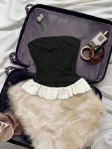 A black strapless tube top with a white ruffled lace hem, styled inside an open black suitcase with a beige faux fur accessory, rings, and a perfume bottle.