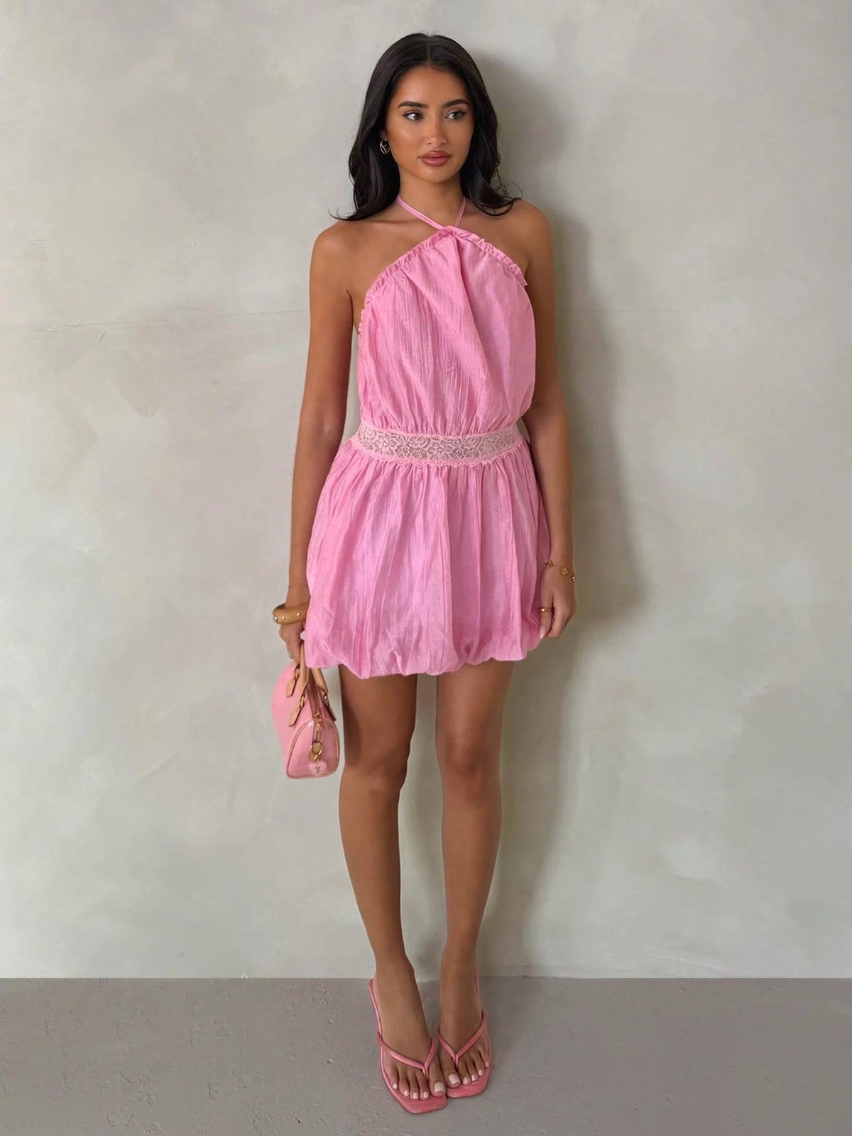 A woman stands against a textured light gray wall wearing a pink halter-neck mini dress with a lace waistband, holding a matching pink handbag and wearing pink sandals.