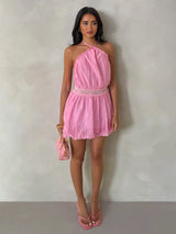 A woman stands against a textured light gray wall wearing a pink halter-neck mini dress with a lace waistband, holding a matching pink handbag and wearing pink sandals.