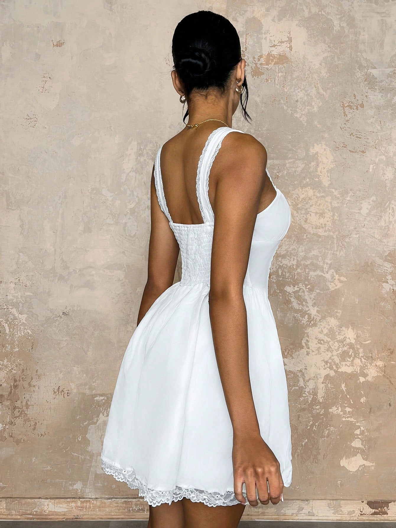 Woman wearing a white dress with lace details against a textured beige wall.