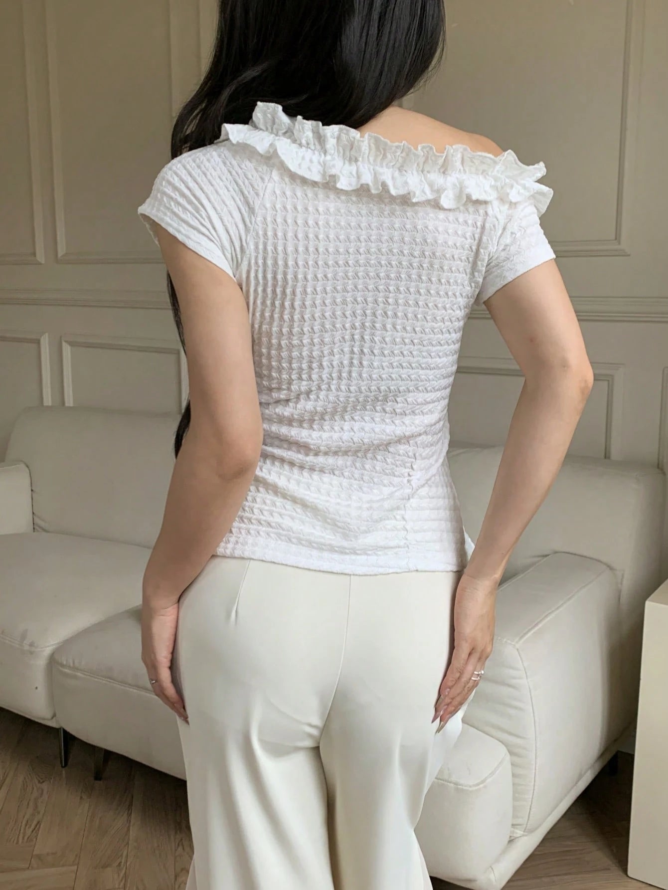Person wearing a white textured top with ruffled shoulders in a room.
