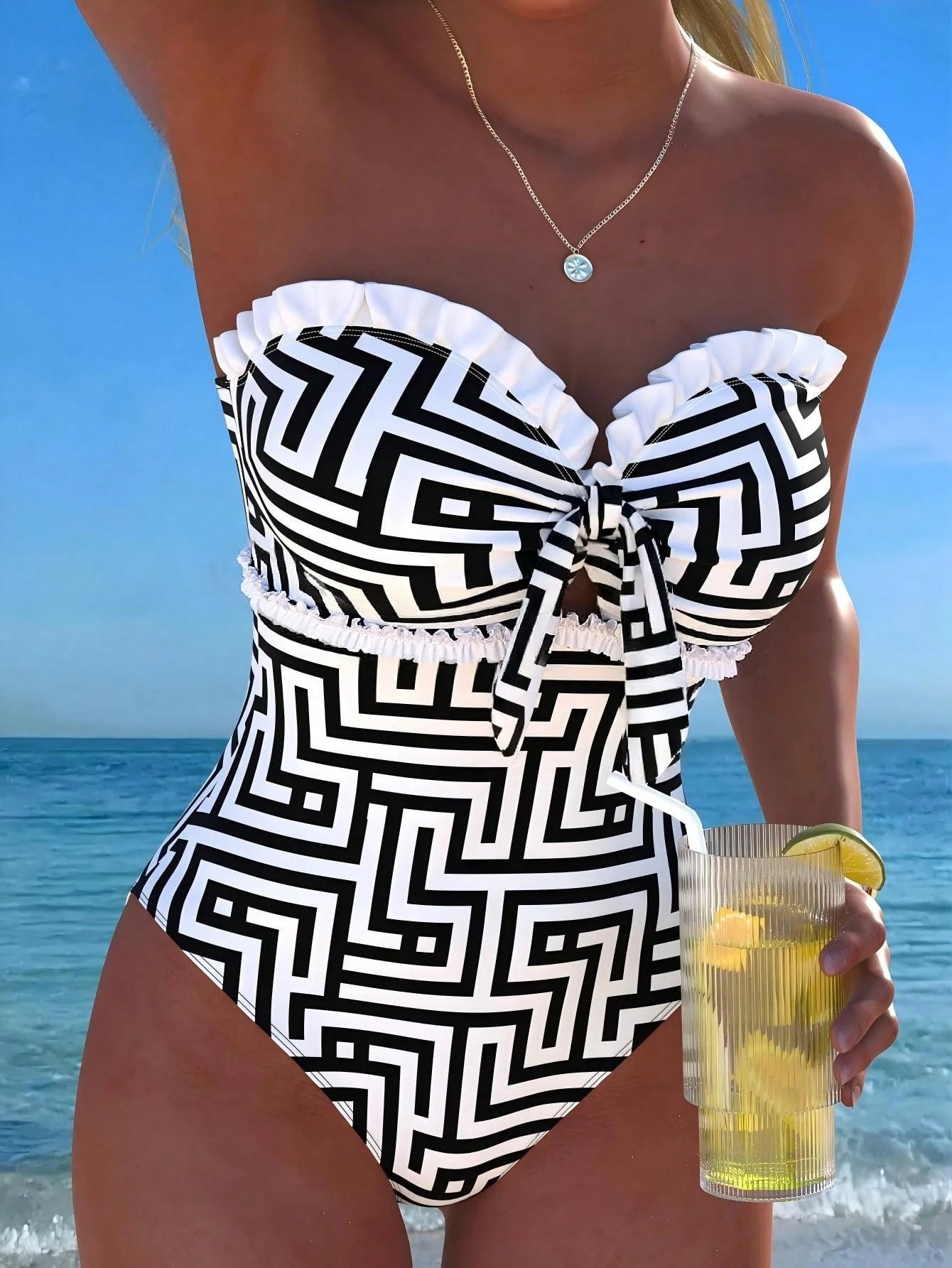 Woman in a black and white patterned swimsuit holding a drink with lemon slices on a beach.